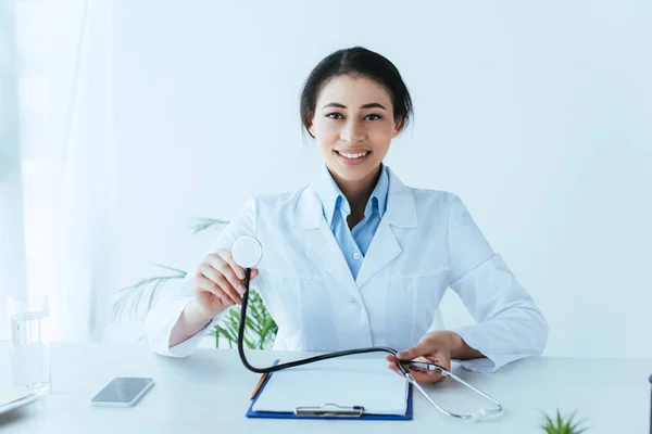 Smiling latin doctor holding stethoscope and smiling at camera while sitting at workplace in clinic — Stock Photo