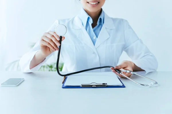 Tiro recortado de jovem médico latino segurando estetoscópio enquanto sentado no local de trabalho — Fotografia de Stock