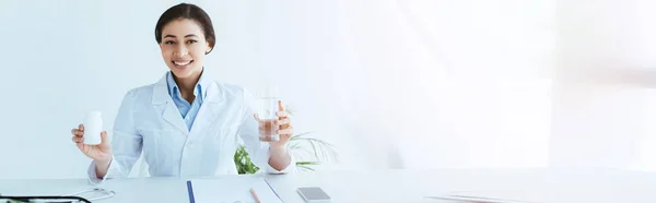 Панорамный снимок улыбающегося латинского врача, держащего стакан воды и контейнер с таблетками — стоковое фото