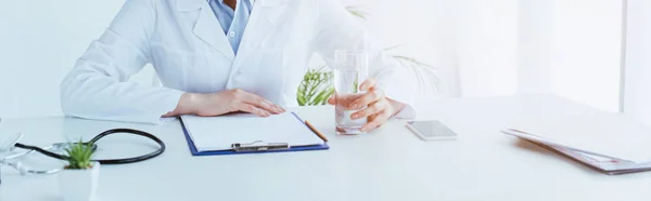 Panoramic shot of doctor holding glass of water while sitting at workplace near clipboard — Stock Photo