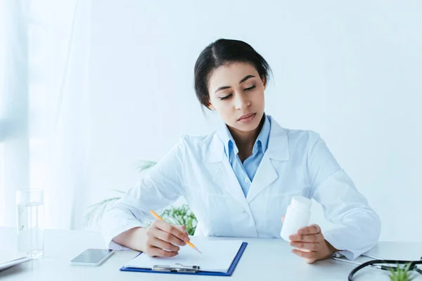 Médico latino atencioso olhando para o recipiente com pílulas enquanto segurando lápis perto da área de transferência — Fotografia de Stock