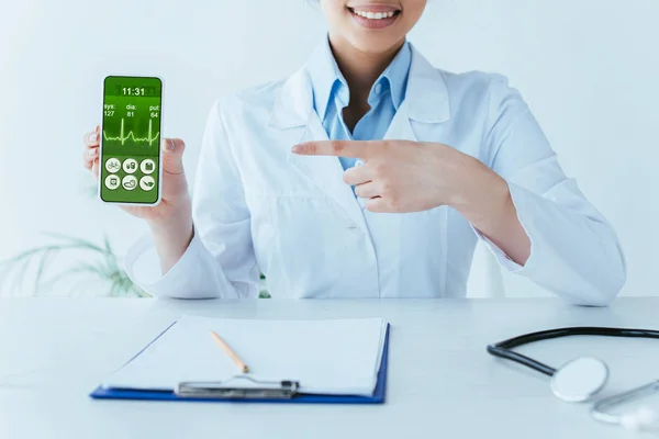 Cropped shot of smiling latin doctor pointing with finger at smartphone with heartbeat rate on screen — Stock Photo