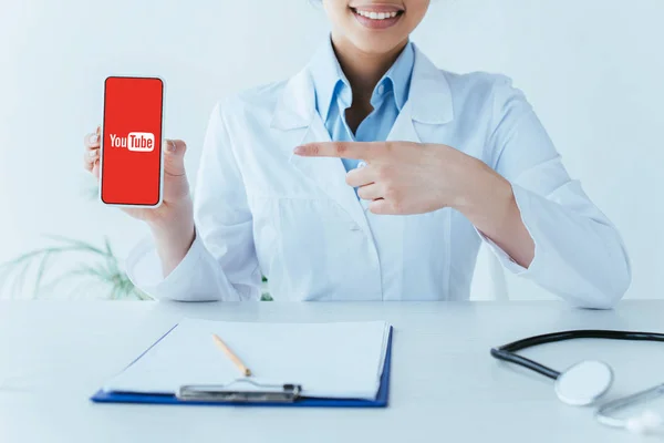 KYIV, UCRANIA - 26 DE ABRIL DE 2019: Vista parcial del médico latino sonriente que señala con el dedo al teléfono inteligente con la aplicación de Youtube en la pantalla . - foto de stock