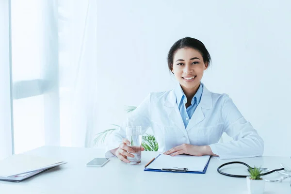 Attractive latin doctor smiling at camera while sitting at workplace with glass of water — Stock Photo