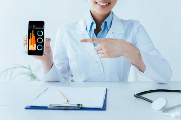 Vista parcial del médico latino sonriente apuntando con el dedo al teléfono inteligente con infografías en la pantalla - foto de stock