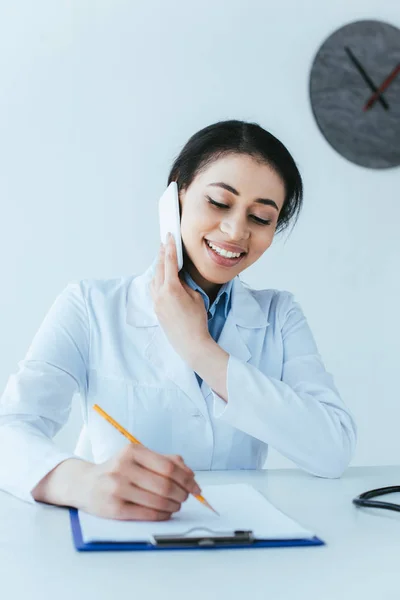 Gai médecin latin écriture diagnostic sur presse-papiers tout en parlant sur smartphone — Photo de stock