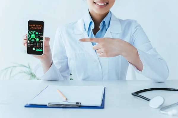 Recortado disparo de sonriente médico latino señalando con el dedo en el teléfono inteligente con aplicación de análisis de marketing en la pantalla - foto de stock