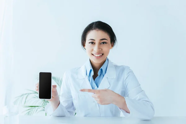 Atractivo médico latino sonriendo a la cámara y apuntando con el dedo al teléfono inteligente con pantalla en blanco - foto de stock