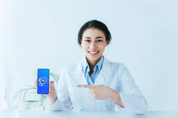 KYIV, UCRANIA - 26 DE ABRIL DE 2019: Médico latino sonriente que señala con el dedo al teléfono inteligente con la aplicación Shazam en la pantalla . - foto de stock