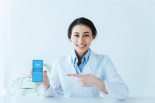 KYIV, UKRAINE - 26 AVRIL 2019 : Joyeux médecin latin pointant du doigt le smartphone avec l'application Skype à l'écran . — Photo de stock
