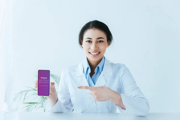 KYIV, UKRAINE - APRIL 26, 2019: Pretty latin doctor pointing with finger at smartphone with Instagram app on screen. — Stock Photo