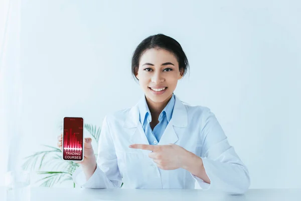 Attractive latin doctor pointing with finger at smartphone with trading courses on screen — Stock Photo