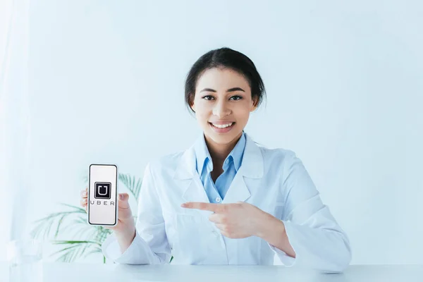 KYIV, UCRANIA - 26 DE ABRIL DE 2019: Hermoso médico latino que señala con el dedo al teléfono inteligente con la aplicación Uber en la pantalla . - foto de stock