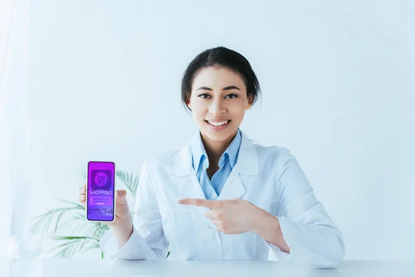 Hermoso médico latino mostrando teléfono inteligente con aplicación de compras en línea en la pantalla - foto de stock