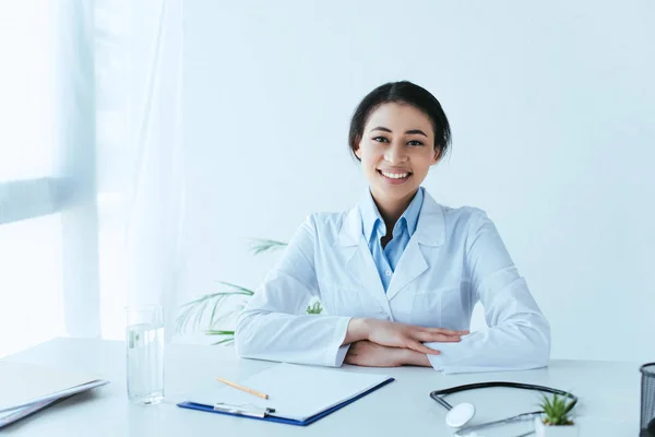 Fröhliche lateinamerikanische Ärztin lächelt in die Kamera, während sie am Arbeitsplatz im Krankenhaus sitzt — Stockfoto