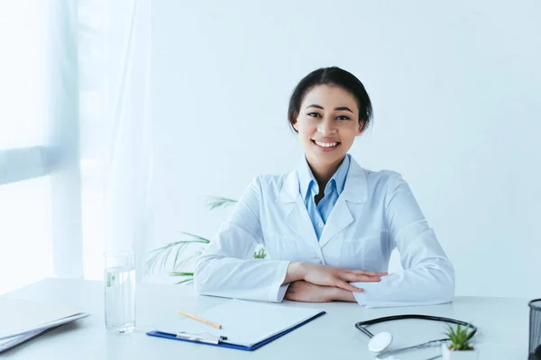 Alegre médico latino sonriendo a la cámara mientras está sentado en el lugar de trabajo en el hospital - foto de stock