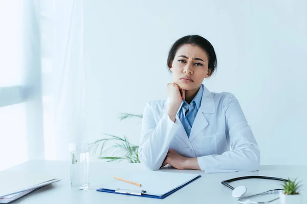 Contrarié médecin latin regardant la caméra tout en étant assis sur le lieu de travail à la clinique — Photo de stock