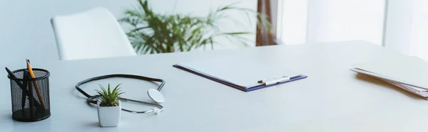 Panoramic shot of clipboard with blank paper, stethoscope and green potted plant on desk in hospital — Stock Photo