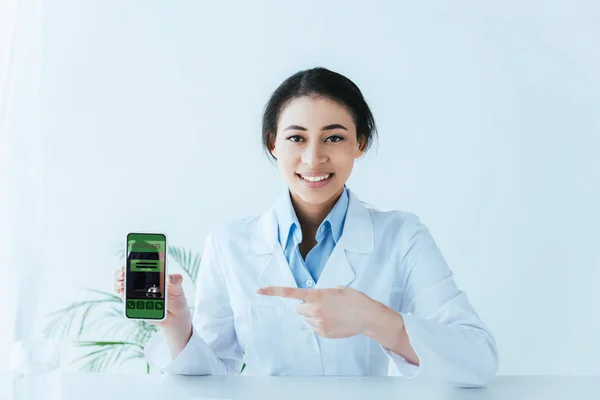 Cheerful latin doctor pointing with finger at smartphone with booking app on screen — Stock Photo