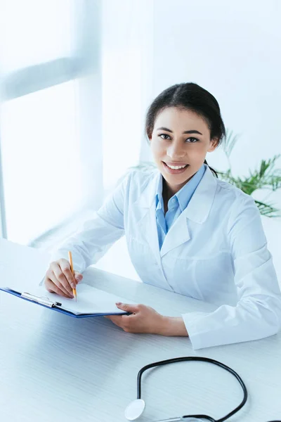 Atractiva doctora latina escribiendo en el portapapeles mientras sonríe a la cámara en la oficina - foto de stock