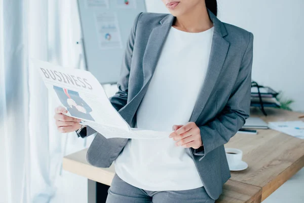 Vista parcial de la mujer de negocios latina leyendo el periódico de negocios mientras está de pie cerca del escritorio - foto de stock