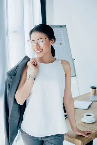 Jolie femme d'affaires latine debout près du lieu de travail et regardant loin dans le bureau — Photo de stock