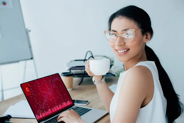 Mujer de negocios latina sonriente usando el ordenador portátil con infografías en la pantalla mientras sostiene la taza de café y sonríe a la cámara - foto de stock