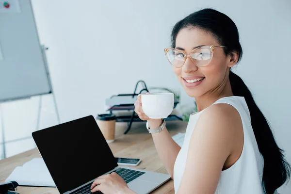 Cheerful latin businesswoman holding coffee cup and using laptop while smiling at camera — Stock Photo