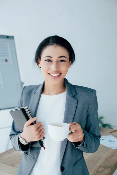 Pretty latin businesswoman holding notebooks and coffee cup while smiling at camera — Stock Photo