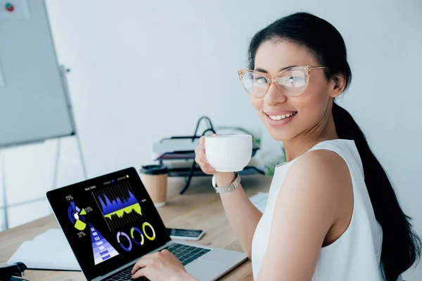 Sorridente latino empresária segurando xícara de café ao usar laptop com gráficos e gráficos na tela — Fotografia de Stock