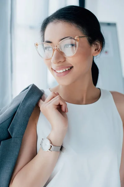 Hermosa mujer de negocios latina en gafas sonriendo y mirando hacia otro lado en la oficina - foto de stock