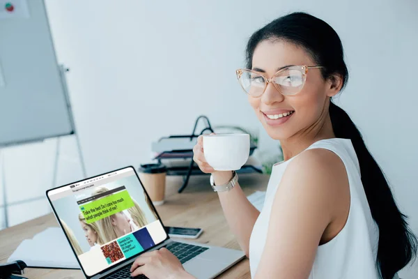 KYIV, UKRAINE - 26 de abril de 2019: Empresária latina atraente segurando xícara de café enquanto usa laptop com o site da BBC Science na tela . — Fotografia de Stock
