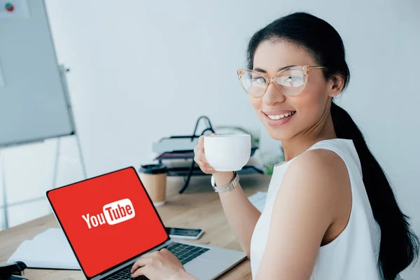 KYIV, UKRAINE - APRIL 26, 2019: Attractive latin businesswoman holding coffee cup while using laptop with Youtube website on screen. — Stock Photo