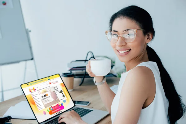 KYIV, UKRAINE - APRIL 26, 2019: Smiling latin businesswoman using laptop with Aliexpress website on screen and holding coffee cup. — Stock Photo