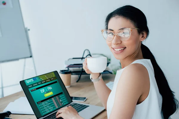 KYIV, UKRAINE - 26 de abril de 2019: Mulher de negócios latina sorridente usando laptop com site Sportsbet na tela enquanto segura xícara de café . — Fotografia de Stock