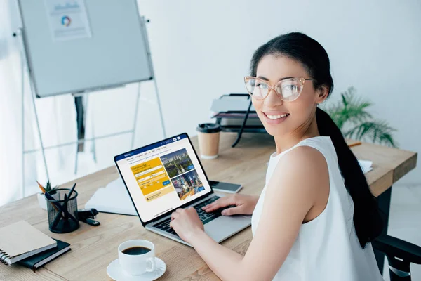 KYIV, UCRANIA - 26 DE ABRIL DE 2019: Bonita mujer de negocios latina en gafas usando el ordenador portátil con el sitio web de reservas en la pantalla mientras sonríe a la cámara . - foto de stock
