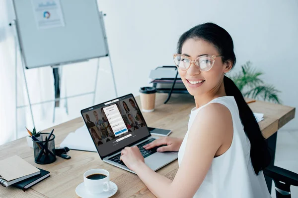 KYIV, UCRANIA - 26 DE ABRIL DE 2019: Mujer de negocios latina sonriente mirando a la cámara mientras usa la computadora portátil con el sitio web Linkedin en la pantalla . - foto de stock