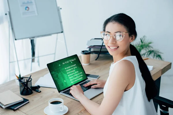 Smiling latin businesswoman looking at camera while using laptop with text information on screen — Stock Photo