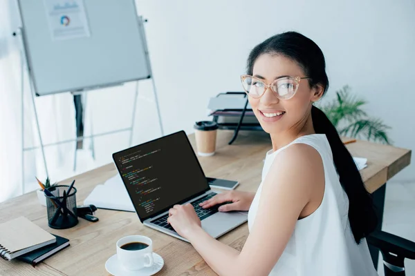 KYIV, UKRAINE - 26 AVRIL 2019 : Femme d'affaires latine attirante souriant à la caméra tout en utilisant un ordinateur portable avec du code html à l'écran . — Photo de stock