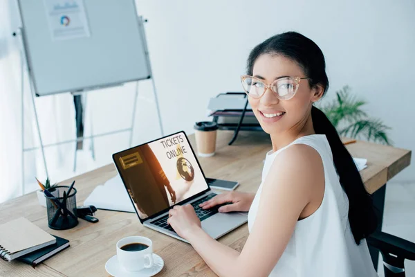 Joyeuse femme d'affaires latine regardant la caméra tout en utilisant un ordinateur portable avec des billets site en ligne à l'écran — Photo de stock