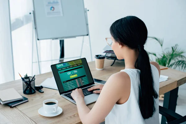KYIV, UCRANIA - 26 DE ABRIL DE 2019: Joven mujer de negocios latina usando la computadora portátil con el sitio web Sportsbet en la pantalla mientras está sentada en el escritorio cerca de la taza de café . - foto de stock