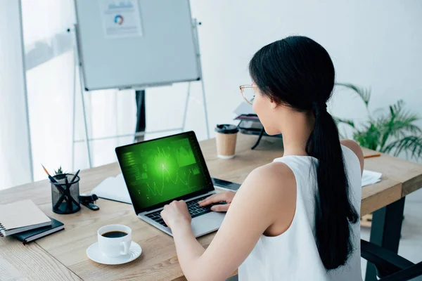 Joven mujer de negocios latina usando portátil con gráficos y gráficos en la pantalla mientras está sentado en el escritorio cerca de la taza de café - foto de stock