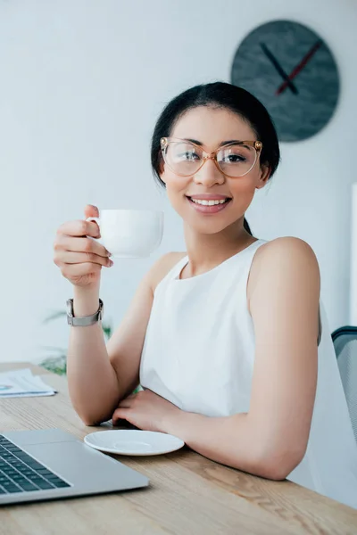 Alegre latina mujer de negocios sosteniendo taza de café mientras sonríe a la cámara - foto de stock