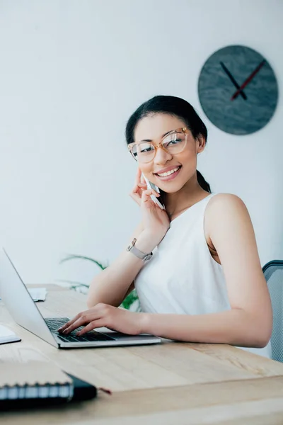 Pretty latin businesswoman talking on smartphone and using laptop at workplace — Stock Photo