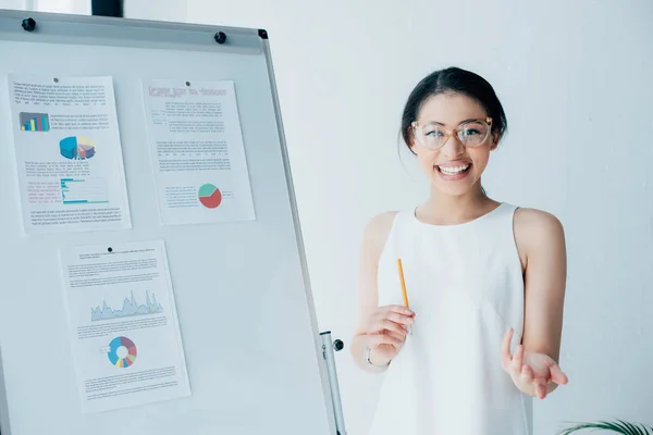 Alegre mujer de negocios haciendo gestos de pie cerca de flipchart con infografías y mirando a la cámara - foto de stock