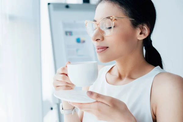 Hermosa mujer de negocios disfrutando del sabor del café mientras sostiene la taza de café con los ojos cerrados - foto de stock