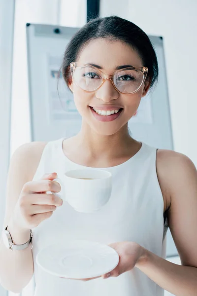 Alegre empresaria en vasos sosteniendo taza de café y sonriendo a la cámara - foto de stock