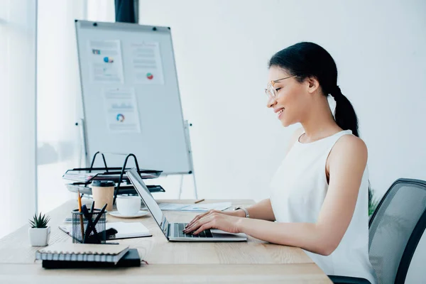 Mujer de negocios latina sonriente escribiendo en el portátil mientras está sentada en el lugar de trabajo en la oficina — Stock Photo