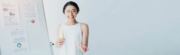 Plan panoramique de femme d'affaires latine joyeuse gestuelle et souriante à la caméra tout en se tenant près de tableau à feuilles mobiles — Photo de stock