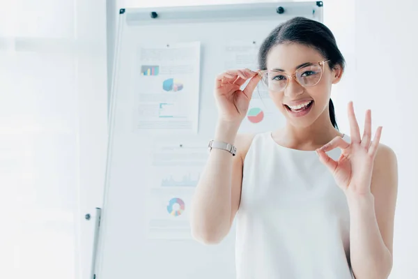 Happy latin businesswoman showing ok gesture while touching glasses and smiling at camera — Stock Photo